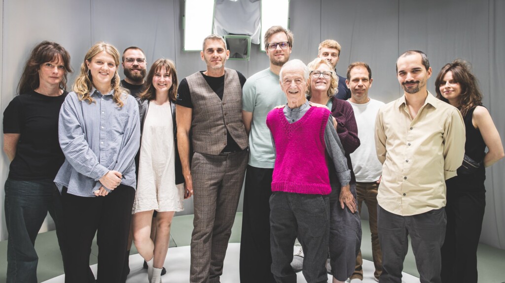 Group photo featuring Holocaust survivor Gidon Lev (center, in a bright pink vest) surrounded by a diverse team of creatives and technicians in a modern volumetric capture studio.